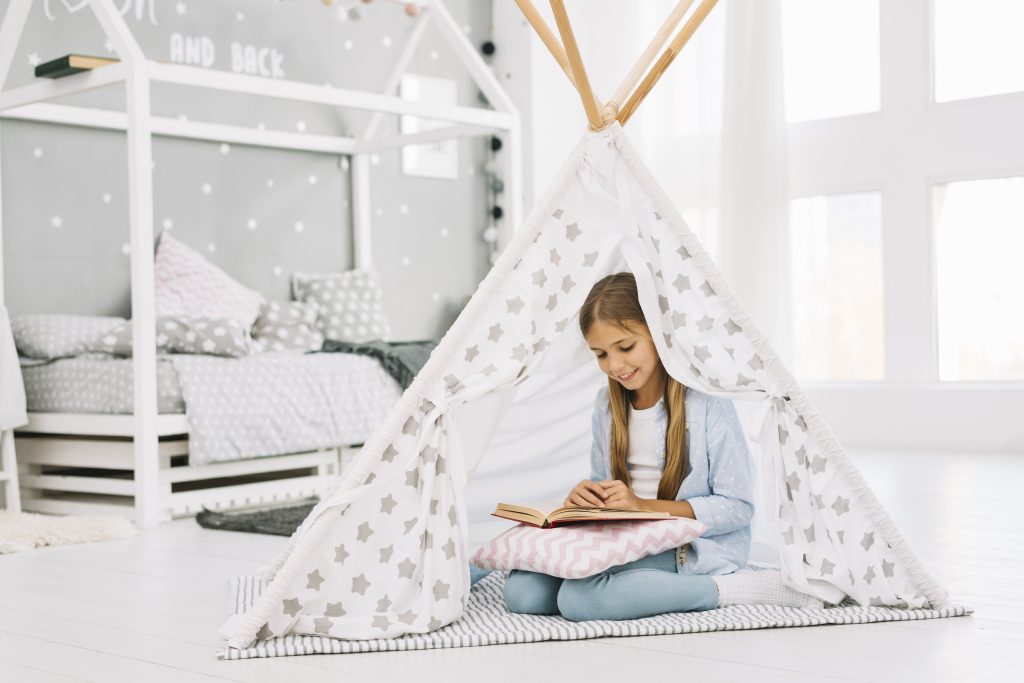 Girl reading a book in a cozy tent with pillows in a beautifully designed kids’ bedroom